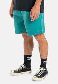 Turkosa bomullsshorts med sidofickor, avslappnad passform och lätt slitna detaljer. Kombineras med svarta sneakers och strumpor med logotyp.