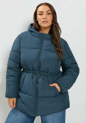 Femme aux longs cheveux foncés portant une doudoune couleur sarcelle avec une capuche et une fermeture éclair, debout avec une main dans une poche sur un fond uni.