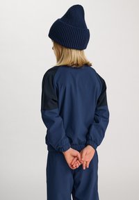 Marinblå jacka med svarta accenter, elastiska muddar och midja. Matcher med en marinblå stickad mössa. Barnets frisyr syns bakifrån.