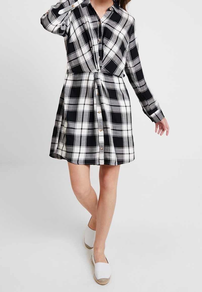 Abito camicia a quadri bianco e nero con chiusura a bottoni, maniche lunghe e dettaglio nodo frontale; realizzato in tessuto intrecciato con una morbida consistenza.