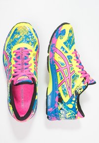 Des chaussures de course aux couleurs vives avec un dessus principalement jaune comportant des motifs éclaboussés bleu et rose, des accents noirs et des lacets roses.