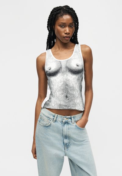 Γυναίκα με πλεξούδες που φοράει ασημένιο τοπ με σχέδιο στο μπροστινό μέρος και ανοιχτό μπλε τζιν, ποζάροντας με το ένα χέρι στην τσέπη.