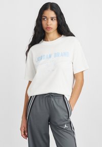 Hosszú sötét hajú nő fehér Jordan Brand pólóban és szürke sportnadrágban, melynek oldalán csíkok vannak, kezével a zsebében, balra néz.