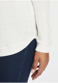 Pulover alb tricotat cu textură ribbed, cu o partea de jos ușor curbată și manșete ajustate, asortat cu blugi de denim de culoare albastru închis. Prim-plan al unei mâini pe material.