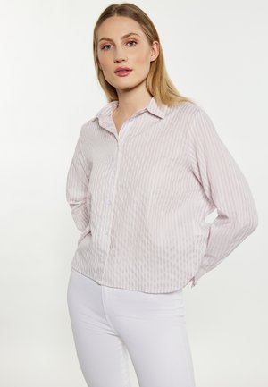 Giovane donna con lunghi capelli biondi indossa una camicia larga a righe viola chiaro con bottoni e pantaloni bianchi su uno sfondo uniforme.