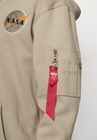 Felpa beige con logo NASA, dotata di tasca con cerniera e etichetta in tessuto rossa. Maniche testurizzate con dettagli di cucitura.