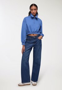 Felpa cropped blu con colletto alto e zip, abbinata a jeans blu a gamba larga e sneakers bianche con dettagli beige.