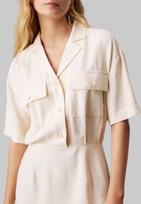 Camicia a maniche corte di colore crema realizzata in tessuto morbido, con colletto a punte, due tasche sul petto e chiusura a bottoni. Tessuto liscio.