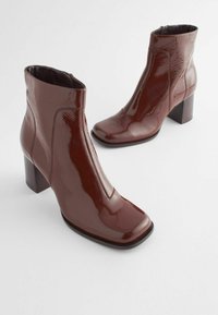 Bottes de cheville en cuir verni bordeaux avec un bout carré, fermeture éclair et talon bloc arrondi, présentant une texture lisse et des coutures minimales.
