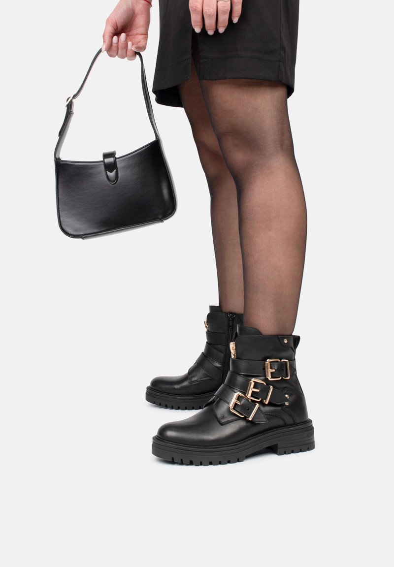 Bottines en cuir noir avec semelles épaisses, ornées de boucles en or et de fermetures éclair ; accompagnées d'un sac à main noir et brillant. Les collants sont transparents.