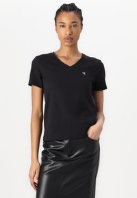 T-shirt nera a maniche corte con scollo a V in cotone, con un piccolo logo CK bianco sul lato sinistro del petto, abbinato a una gonna di pelle nera.