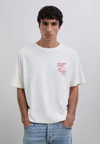 Młody mężczyzna noszący białą koszulkę z czerwoną grafiką kostek do gry i wiśni oraz napisem "SCALPERS GAME" na lewej górnej części klatki piersiowej, stojący na jednolitym tle.