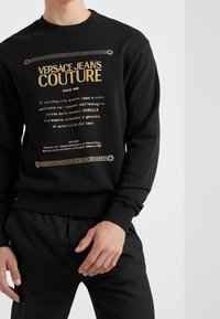 Sudadera negra con texto dorado y diseño de cadena, fabricada en material suave, con puños y dobladillo acanalados. Estilo minimalista con un branding sutil.