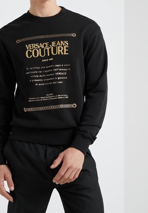 Black sweatshirt featuring gold text and chain design, made of soft material, with ribbed cuffs and hem. Minimalist style with subtle branding.