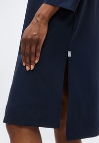 Σκούρο χέρι με ουδέτερο βερνίκι νυχιών να ακουμπάει σε φόρεμα ναυτικού μπλε μήκους μέχρι το γόνατο με πλαϊνή σχισμή και ορατή ταμπέλα με το λογότυπο.