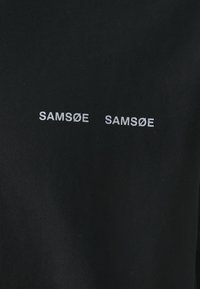 Svart stoff med den broderte teksten "SAM SøE" i hvitt, vist to ganger i en enkel, ren skrifttype. Jevn tekstur, minimal design.