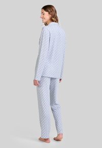 Mujer de pie descalza, vestida con una blusa de manga larga azul claro y pantalones con un pequeño patrón de hojas blancas, sonriendo y mirando de lado.