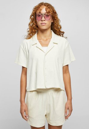 Chemise à manches courtes beige clair à boutons avec une coupe décontractée, associée à un short assorti. Le tissu semble doux avec une texture subtile.