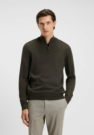 Khaki zip-neck sweater with long sleeves, ribbed cuffs, and hem. Worn with light gray trousers, featuring a smooth texture and fitted design.