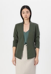 Olijfgroene blazer met een gestructureerd ontwerp, open voorkant en driekwartmouwen, gecombineerd met een turquoise top en een beige geribbelde rok.
