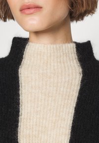 Gros plan du bas du visage et du cou portant un pull beige en maille côtelée sous un manteau noir duveteux.