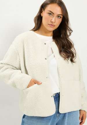 Femme aux longs cheveux bruns porte une veste sherpa blanche par-dessus une chemise blanche et un jean bleu, la main dans la poche de la veste.