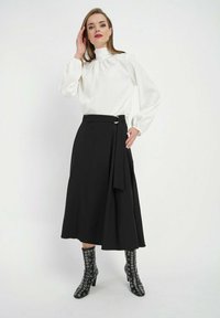 Biała bluzka z teksturą w kropki i wysokim kołnierzykiem, zestawiona z czarną spódnicą typu wrap z paskiem oraz czarnymi kozakami do kolan z metalowymi oczkami.