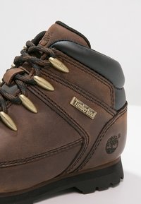 Brązowy skórzany but trekkingowy z złotymi oczkami, czarnym, wyściełanym kołnierzem oraz charakterystycznym logo Timberland. Teksturowana powierzchnia z detalami szwów.