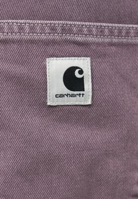 Carhartt címke világos, textúrájú lila anyagon, fehér címkével, fekete logóval és fekete felirattal. Látható varrási részletek.