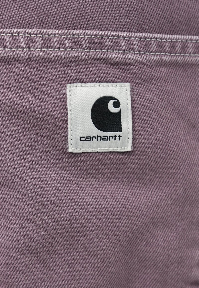 Carhartt-Label auf weichem, strukturiertem lila Stoff, mit einem weißen Etikett, das ein schwarzes Logo und schwarze Schriftzüge zeigt. Sichtbare Nähte.