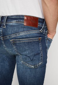 Jeans de mezclilla azul oscuro con un corte ajustado, que presentan un parche de cuero marrón con la marca "Pepe Jeans", costuras en contraste y dos bolsillos traseros.