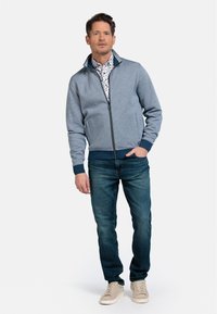 Grijze zip-up jas met contrasterende marineblauwe kraag en manchetten, gedragen met een witte, patroonshirt en blauwe spijkerbroek. Tan sneakers maken de look af.