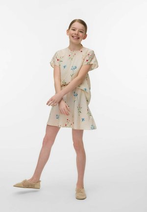 Uśmiechnięta dziewczyna w beżowej sukience w kwiaty i beżowych plecionych wsuwanych butach stoi z rozstawionymi nogami i skrzyżowanymi ramionami na przodzie.