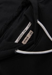 Černá bavlněná mikina s bílým šňůrkami. Má dvouvrstvou kapuci a štítky viditelné na límci. Hladký povrch látky.