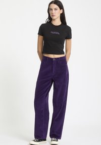 T-shirt noir court avec texte violet associé à un pantalon en velours côtelé violet taille haute et coupe large. Doté de poches avant et d'une fermeture à bouton.