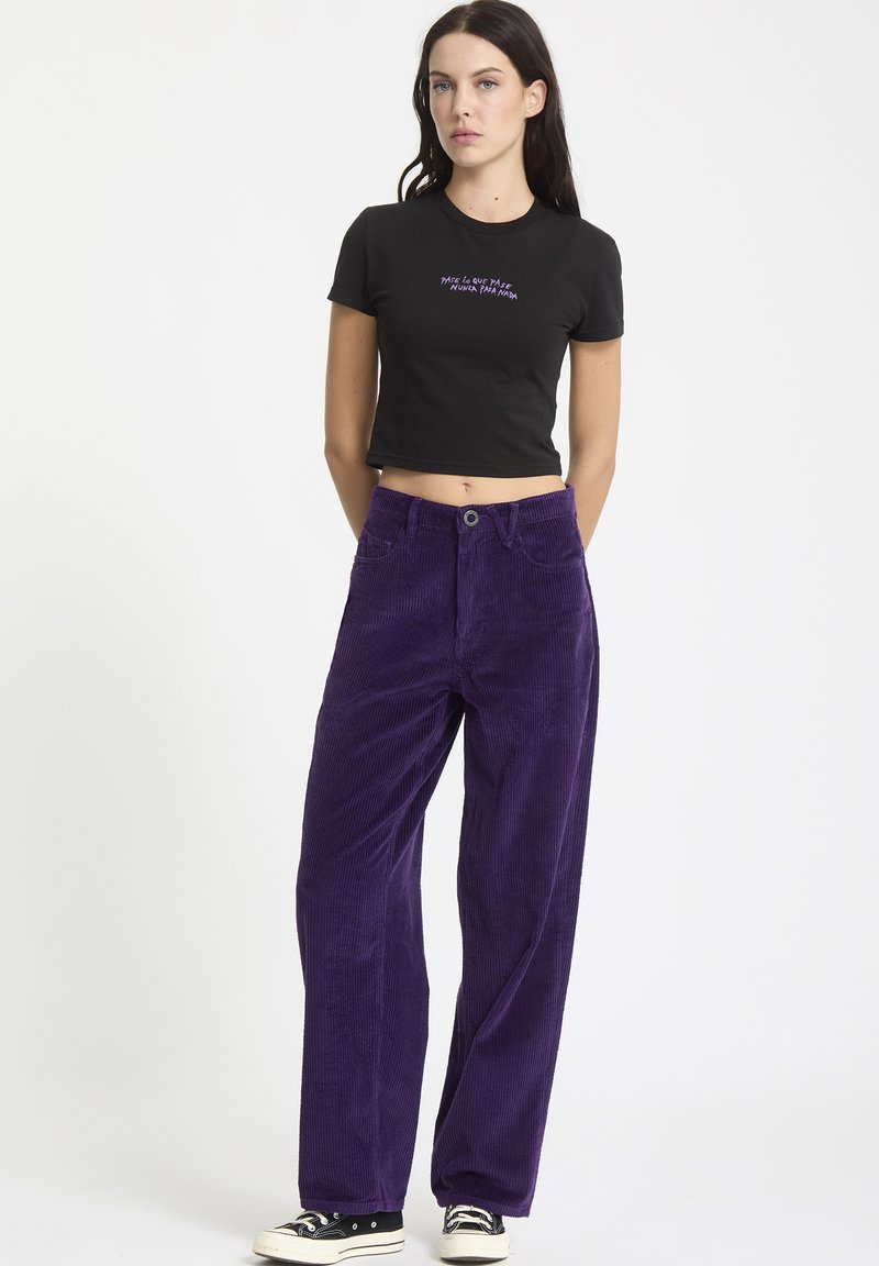 Sort cropped t-shirt med lilla tekst, parret med højtaljede, vide lilla kordfløjl bukser. Har frontlommer og knaplukning.