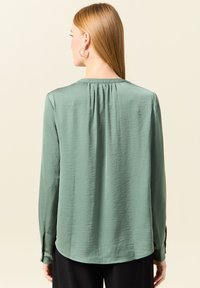 Groene blouse met lange mouwen, een verzamelde rugnaad, zachte textuur en een ronde zoom. Voorzien van knoopmanchetten en een ontspannen pasvorm.