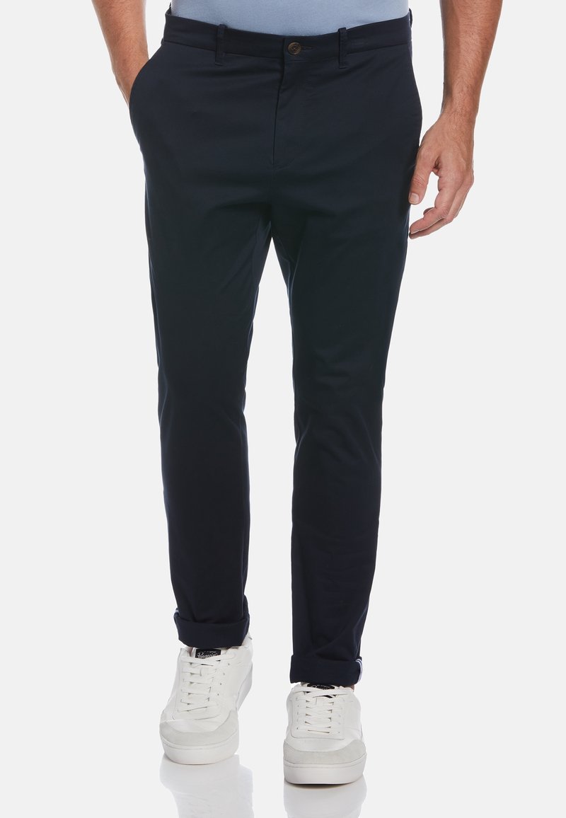 Homme portant un pantalon slim bleu marine foncé avec les revers roulés et des baskets blanches, debout avec une main dans la poche.