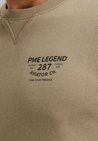 Khaki sweatshirt met geribbelde kraag. Bevat donkere tekst met "PME LEGEND" en vluchtgegevens. Zacht, textuurstof met een getailleerd ontwerp.
