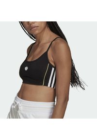 Kvinna som bär en svart sport-bh med vita ränder och gul piping, i kombination med vita byxor med dragsko, visad från axlarna till midjan.