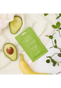 Närande hårbottenpack i grön förpackning, med illustrationer av avokado och banan. Två avokadohalvor och en banan på en krämfärgad bakgrund.