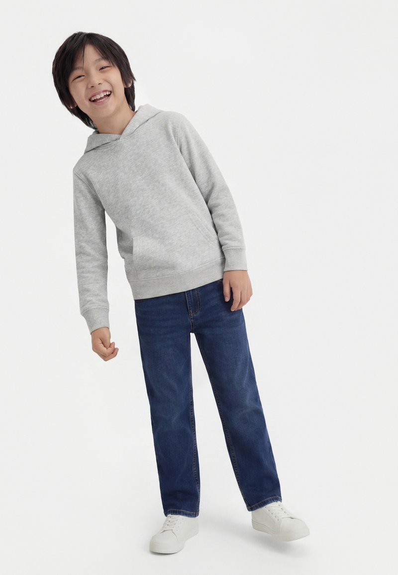 Enfant souriant portant un sweat à capuche gris clair, un jean bleu et des baskets blanches, se tenant légèrement penché contre un fond blanc uni.