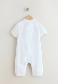 Combinaison courte à manches courtes blanche avec un col rond et des ouvertures élastiques aux jambes. Présente des coutures décoratives le long des manches. Suspendue à un cintre en bois.