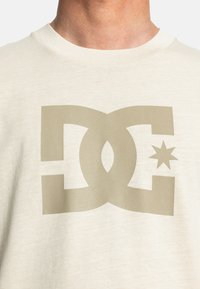 Beige tričko s velkým, tlumeným béžovým logem DC a designem hvězdy umístěným na hrudi. Měkká textura s kulatým výstřihem.