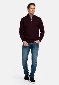 Un pull à col zippé bourgogne avec des poignets et un ourlet côtelés, associé à un jean bleu clair et des sneakers en daim noir, illustrant le confort décontracté.