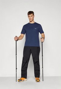 Tmavě modré tričko s krátkým rukávem a přední kapsou, kombinované s černými kalhotami a oranžovými turistickými botami, držící trekové hole.