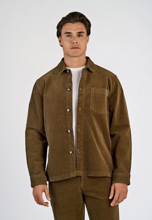 Mand med brun kordfløjls-knap-op skjorte og matchende bukser iført en hvid t-shirt nedenunder, stående mod en ensfarvet baggrund.