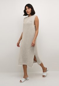 Strickärmloses Kleid in hellem Beige, mit einem offenen Webmuster, seitlichen Schlitzen und gerippten Akzenten, kombiniert mit weißen Absatzmules.