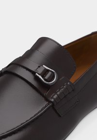 Mocassin en cuir brun foncé avec un détail de boucle en métal et des coutures apparentes sur la tige, présenté en vue rapprochée et angulée.