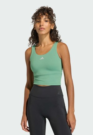 Frau mit lockigem Haar, die ein grünes ärmelloses Crop-Top und hoch taillierte schwarze Leggings vor einem einfarbigen Hintergrund trägt.